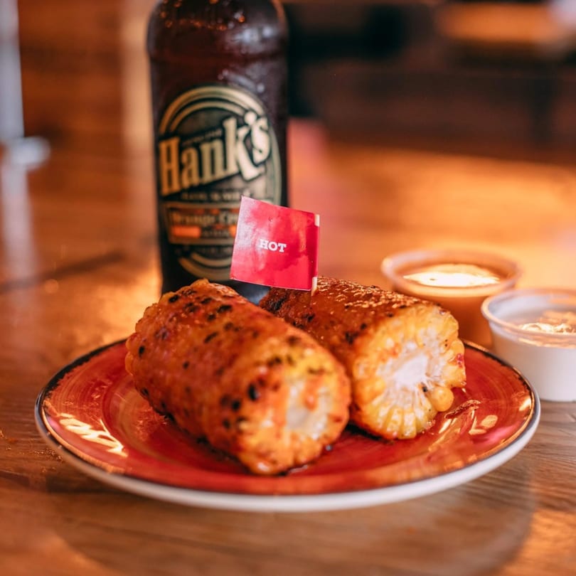 Two grilled corn on the cob with a "HOT" flag, a bottle, and sauces on a wooden table.