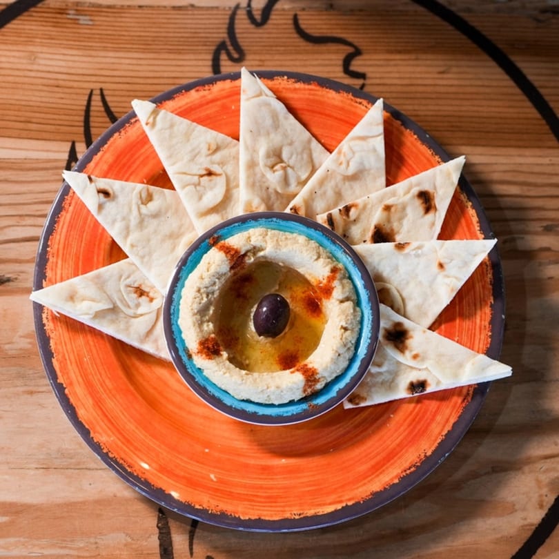 Hummus with an olive and paprika, surrounded by pita bread triangles on an orange plate.