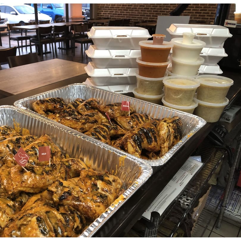 Two foil trays of peri-peri chicken with sauce, takeout containers, and plastic cups of sauces.