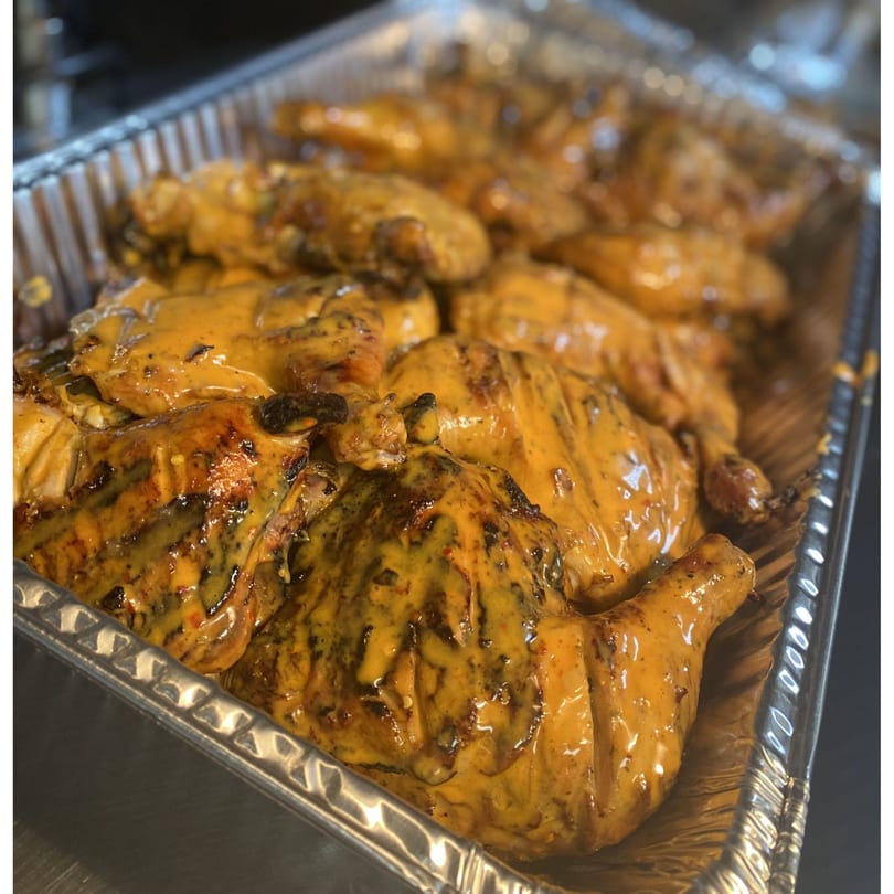 A foil tray filled with many pieces of golden-brown peri-peri chicken covered in a creamy orange sauce.