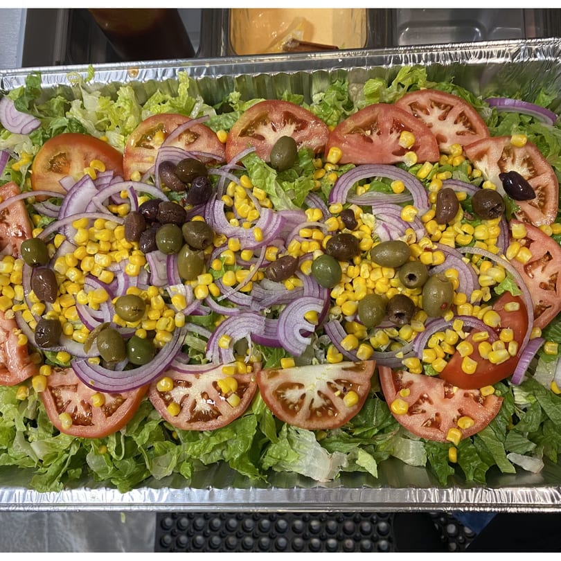 A large catering tray of fresh green lettuce, sliced tomatoes, red onions, corn, and olives.