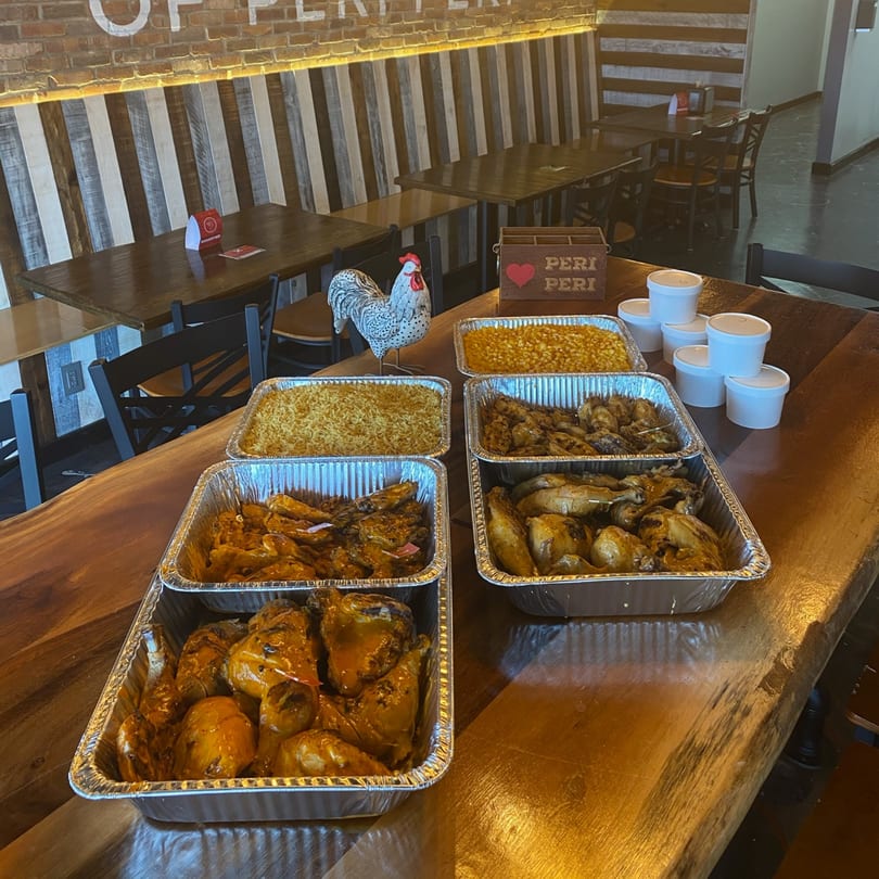 Catering spread of peri-peri chicken, rice, and corn in foil pans on a wooden table.