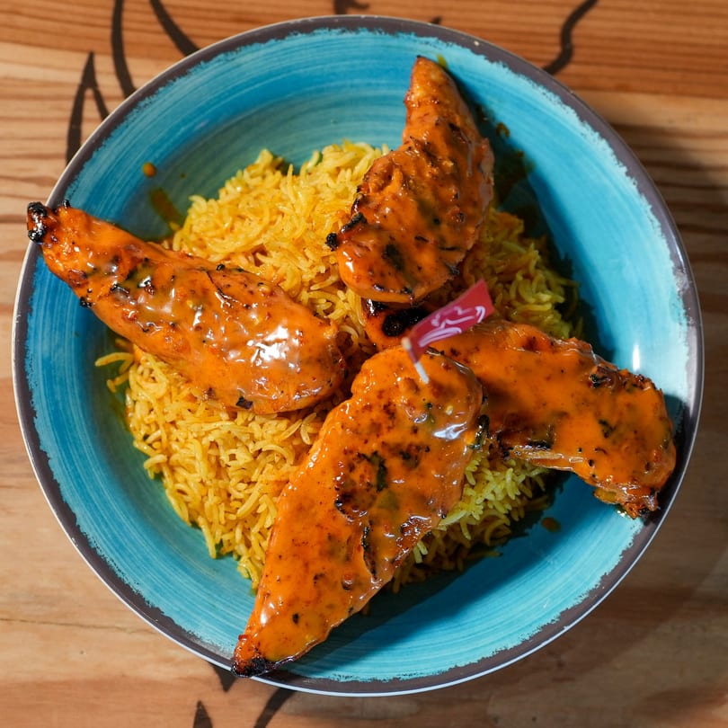 Four peri-peri chicken tenders with rice in a blue bowl on a wooden table.