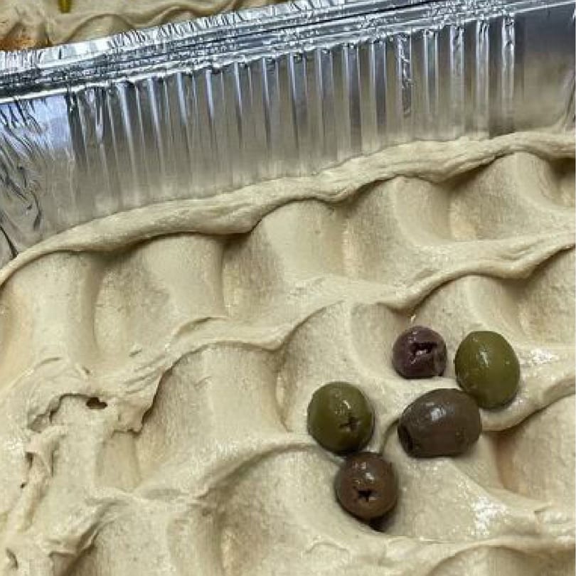 Hummus with green and black olives in a foil container, ready for The Port of Peri Peri customers.