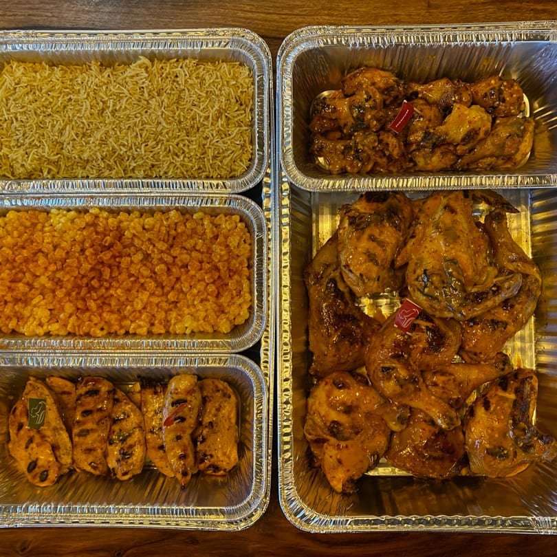 Multiple foil trays filled with peri-peri chicken, rice, and corn, ready for catering.