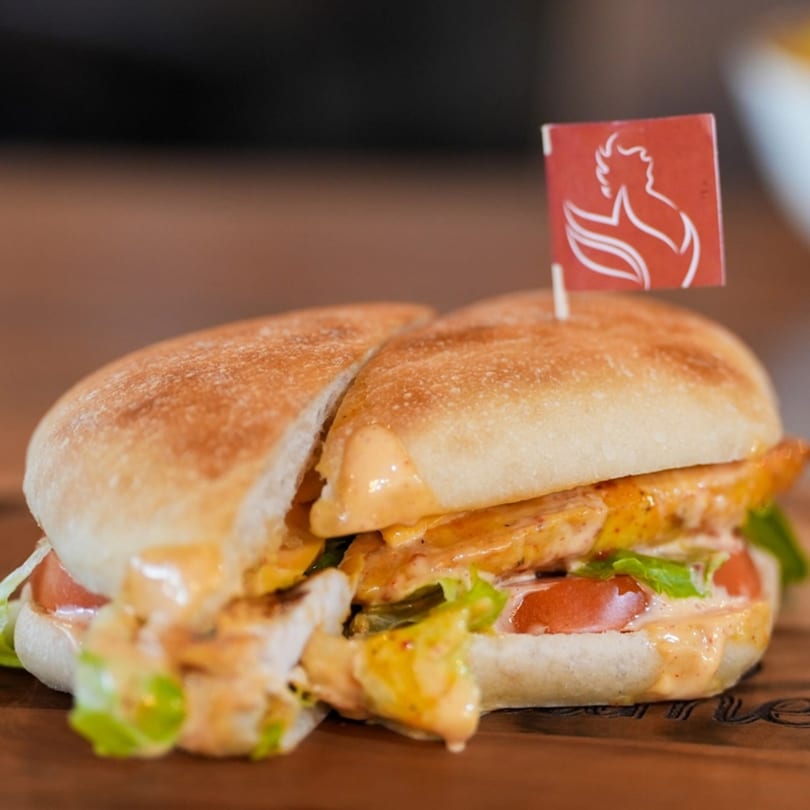 A peri-peri chicken sandwich, cut in half, with lettuce, tomatoes, and sauce on a wooden board.